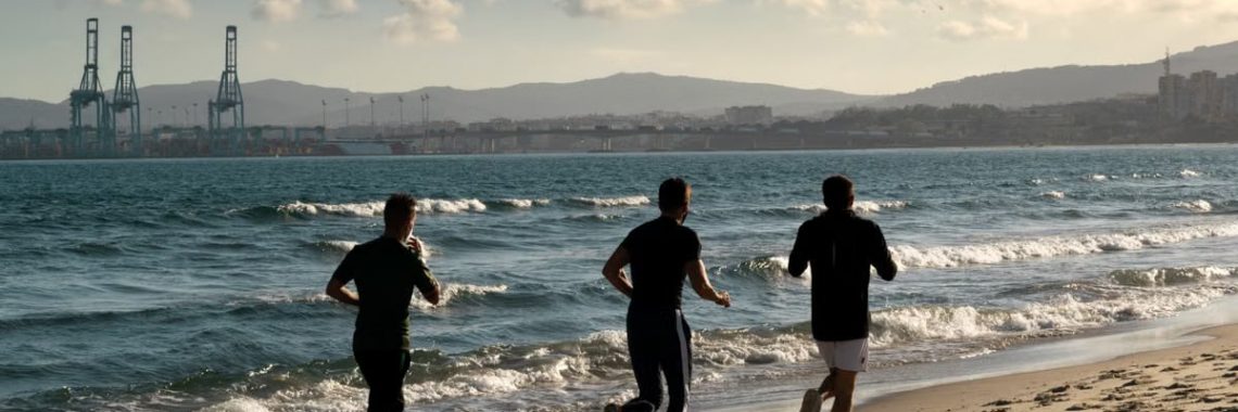 Three men jogging along a scenic beach, embracing a healthy lifestyle under a clear sky.